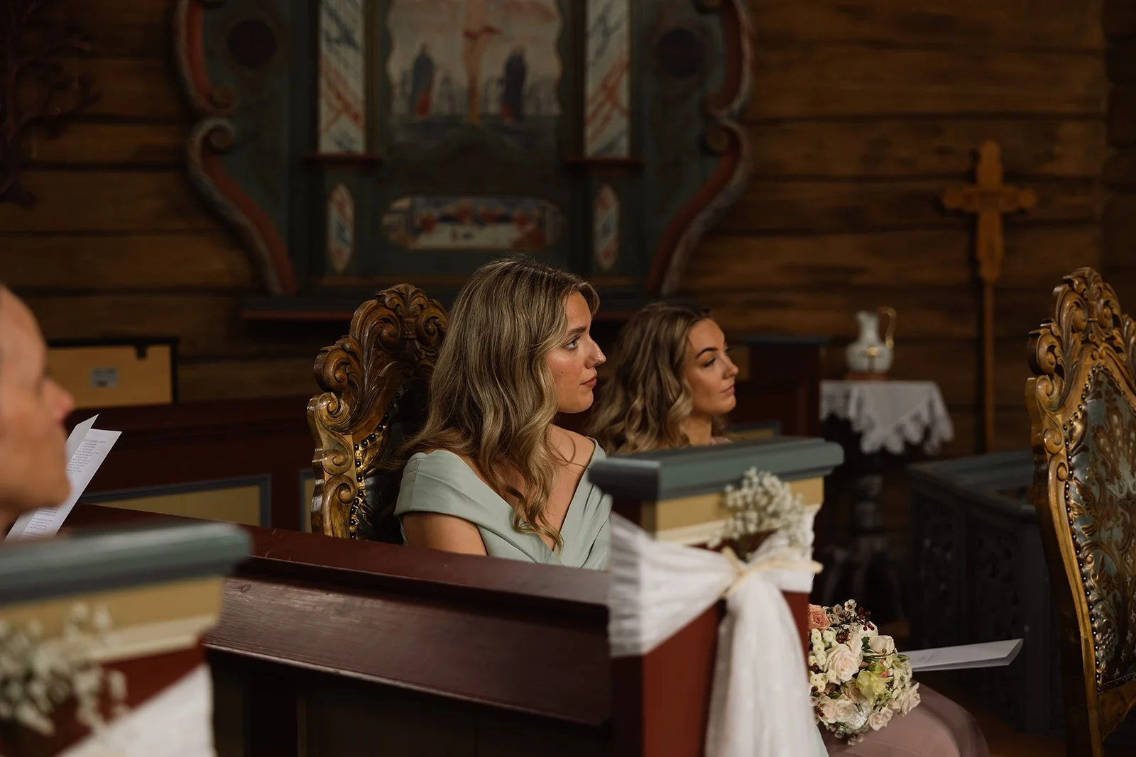To kvinner som sitter i en kirke, en av dem holder en bunad med blomsterbukett. Denne scenen ser ut til å være fra en bryllupseremoni.