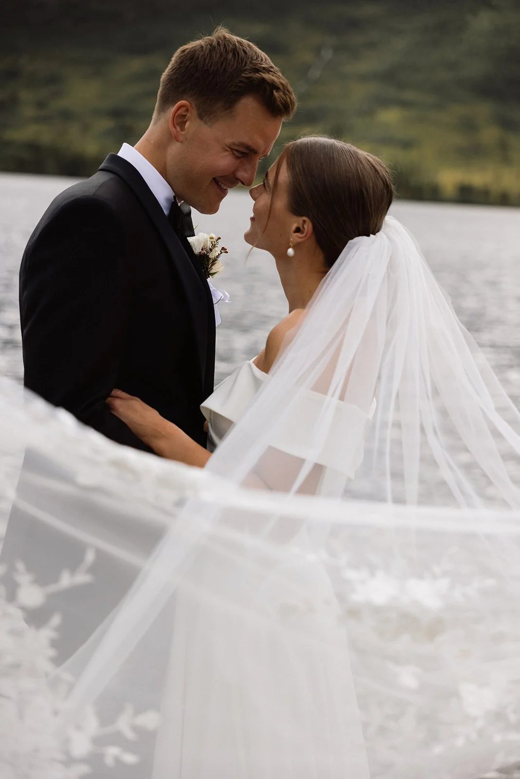 Et brudepar står nær hverandre, smilende og har pannen mot panna foran en naturskjønn bakgrunn med vann og fjell.