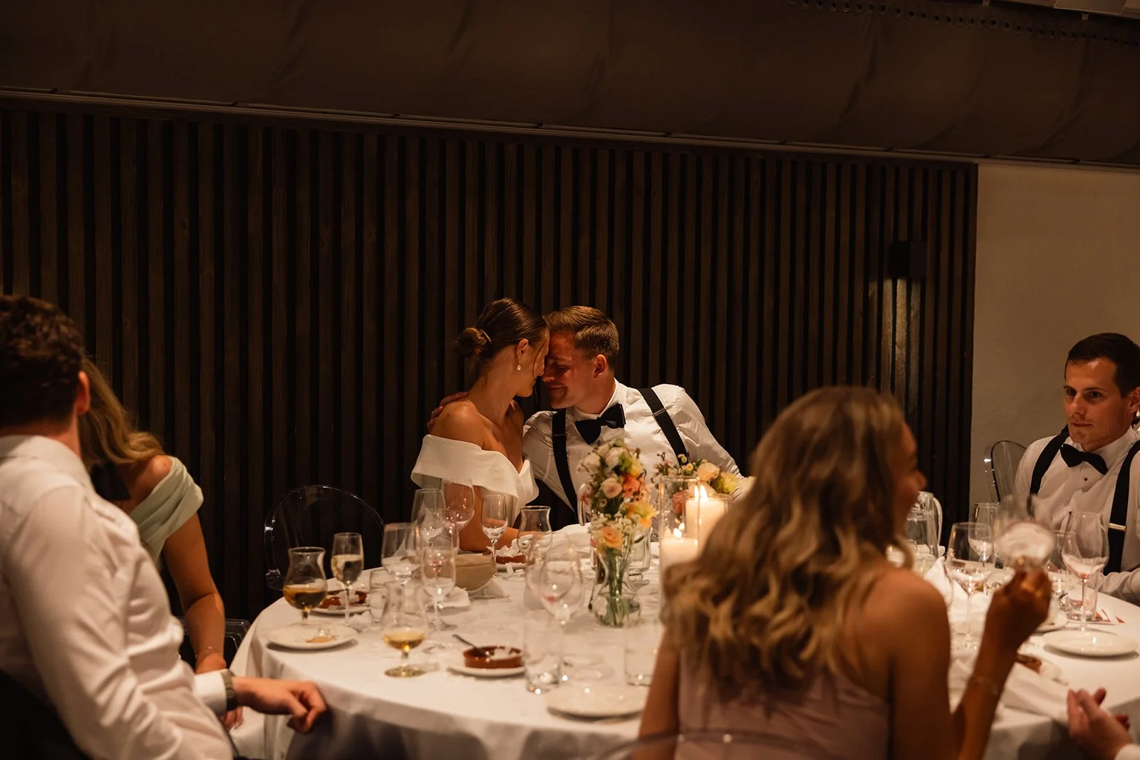 En gruppe mennesker i formelle klær som spiser og prater ved et rundt bord med lysestaker og blomster. To personer nærmest midten har pannen mot pannen i en intim gest.
