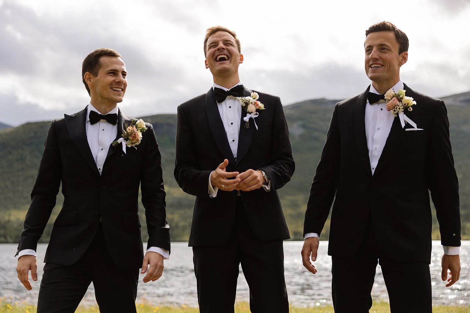 Tre menn i formelle dresser med blomster om brystene, som ler og smiler ved en innsjø med fjell i bakgrunnen.