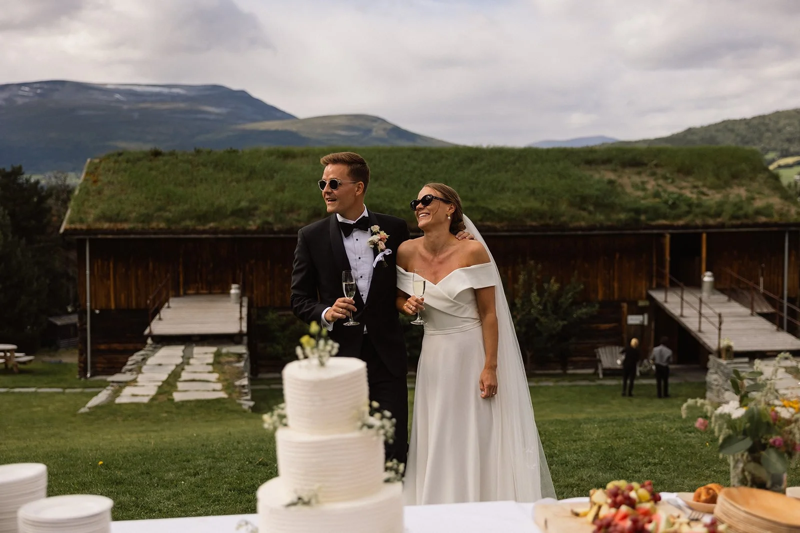 Et brudepar smiler og holder champagneglass under utendørs bryllupsseremoni med et hvitt, lagdelt bryllupskake og matretter foran.