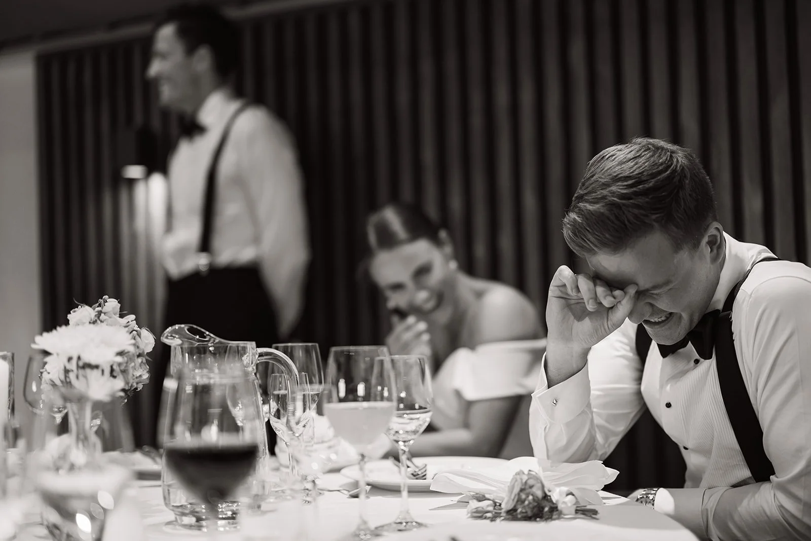 Mann som ler og dekker øyet sitt med hånden, kvinner som ler i bakgrunnen i en formell bankettsetting.