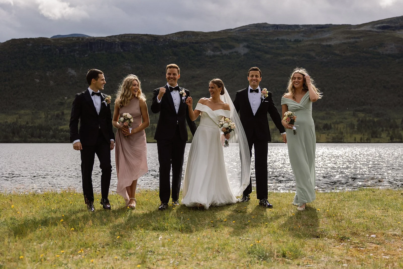 Bryllupsfeiring med syv personer, tre menn i svart dress, tre kvinner i fargede kjoler og en brud i hvit kjole og slør ved en innsjø med åser i bakgrunnen.