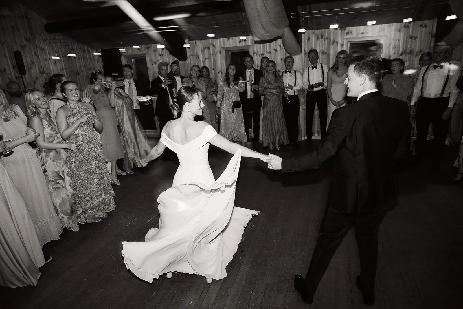 Sort-hvitt bilde av et bryllupsdansende ektepar omgitt av venner i et trehus innendørs.