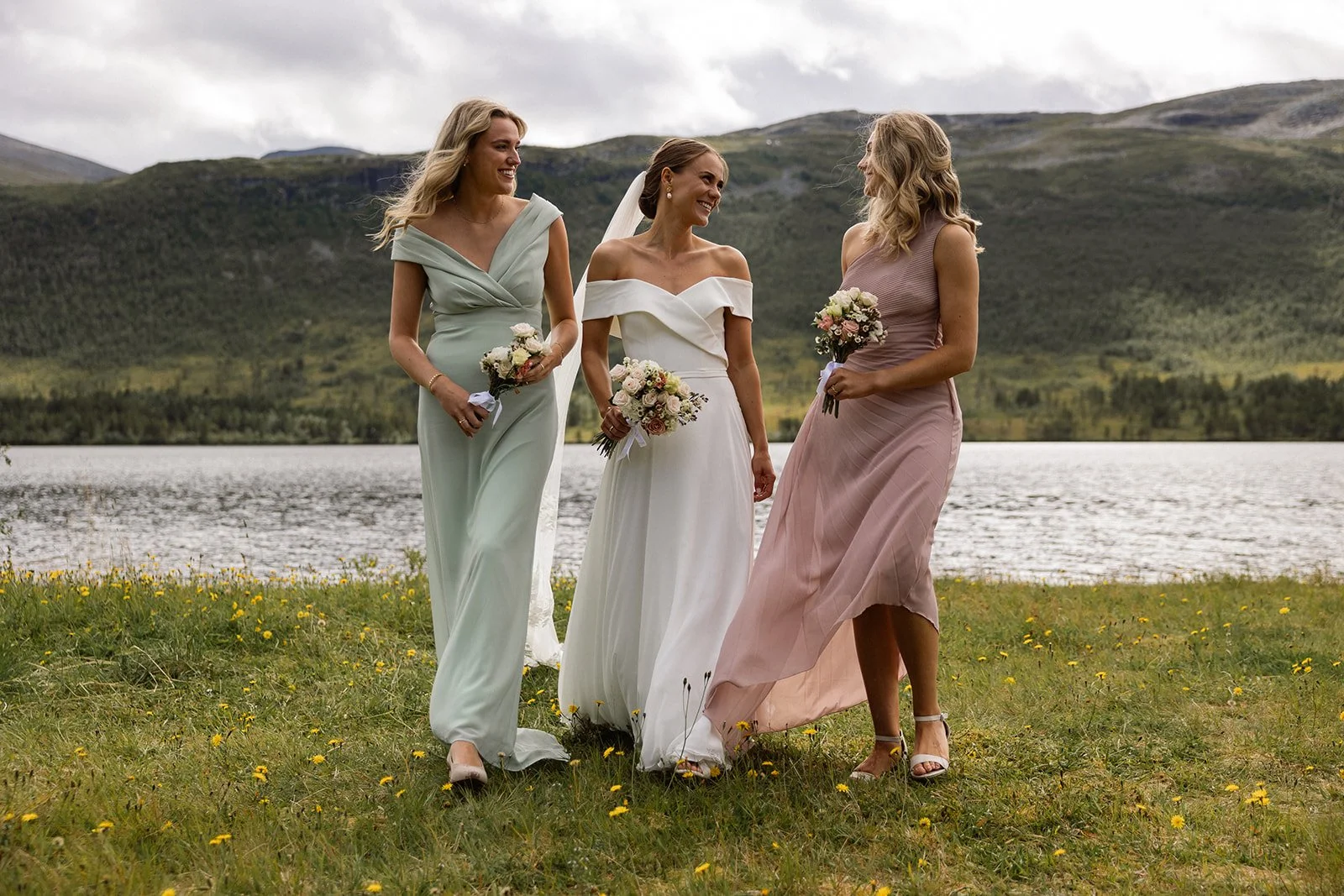 Tre kvinner i brudekjoler som går sammen ved innsjø omgitt av fjell