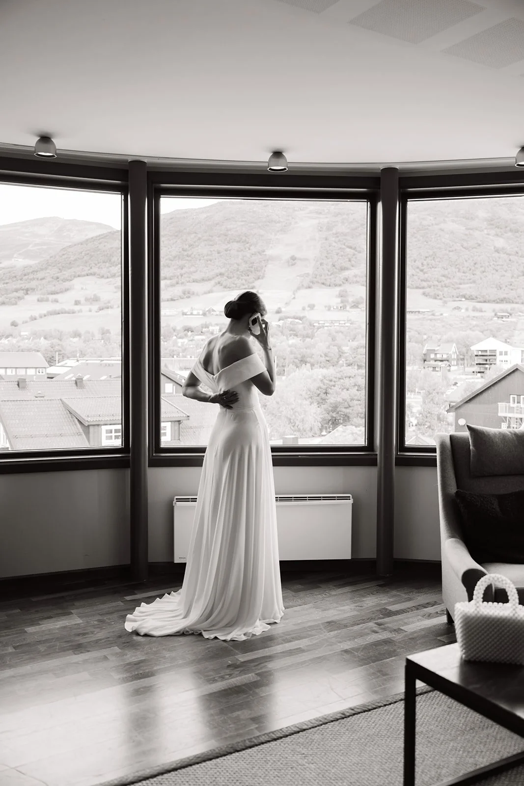 En kvinne i en lang, hvit kjole står ved et vindu og snakker i telefonen, med utsikt over en landsby og fjell i bakgrunnen.