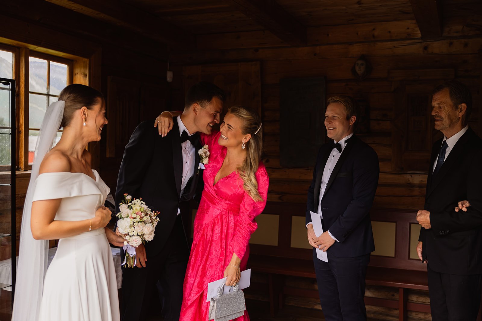 Et brudepar iogat i et rom med tre menn, hvorav to er i smoking, og en kvinne i en lang, rosa kjole. De ler og smiler, og det er klart at det er et lykkelig øyeblikk, sannsynligvis under en bryllupsseremoni.