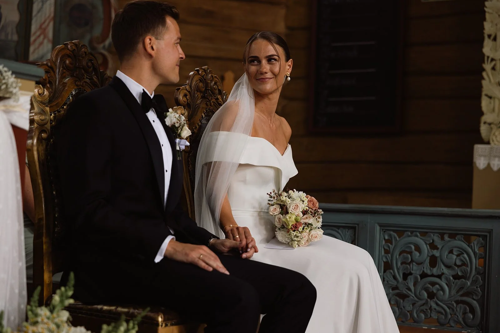 En brud og en brudgom som sitter på en tronlignende stol under et bryllup, med bruden i en hvit kjole og brudgommen i smoking