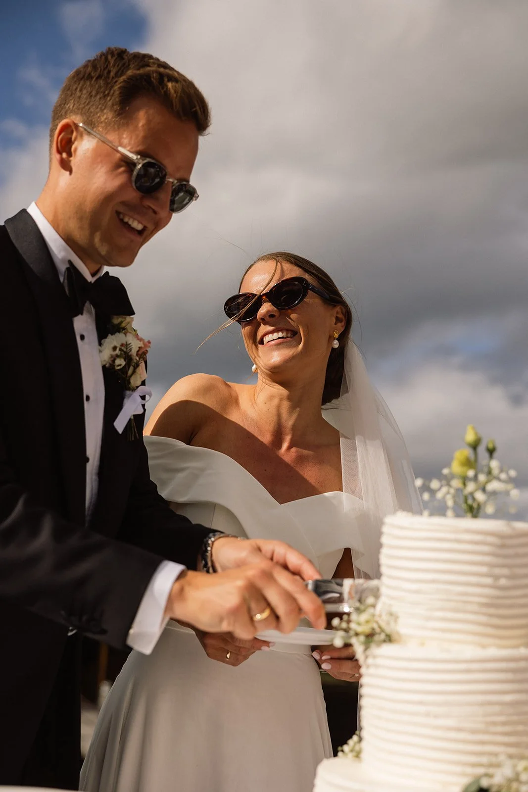 En brud og en brudgom, begge med solbriller, smiler mens de skjærer i en bryllengåte foran en hvit brylliskake med blomster.