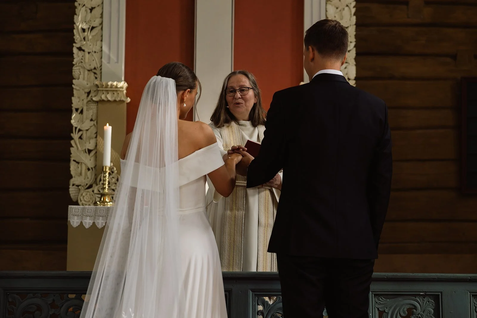 En brud og en brudgom holder hender under vielsen foran en prest i en kirke.