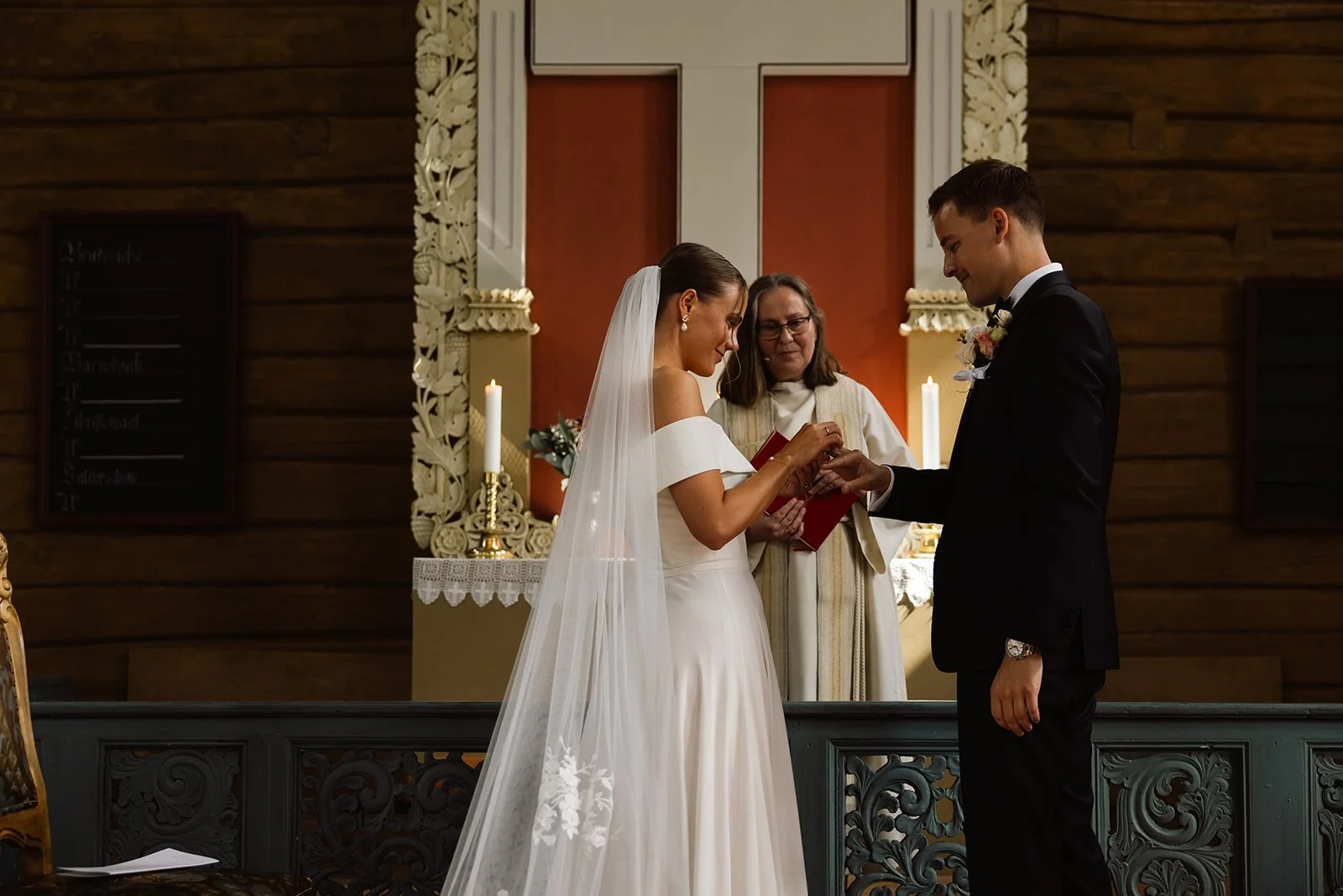 Et brudepar står foran en prest i en kirke under vielsen, med lysende stearinlys i bakgrunnen.