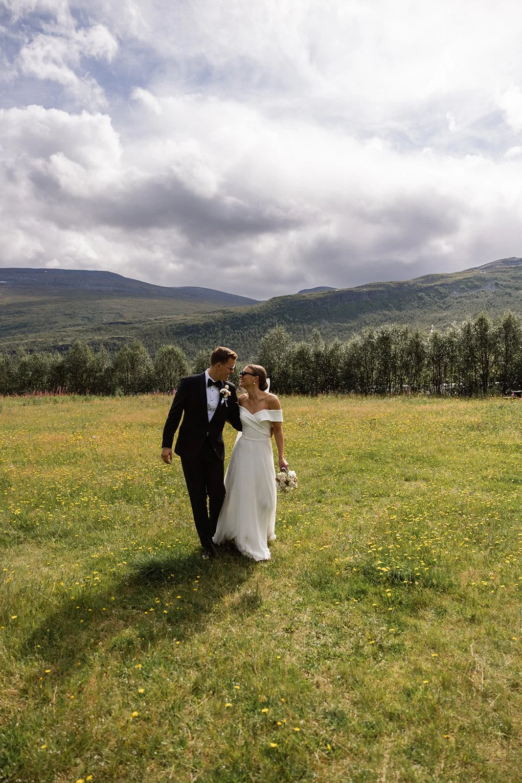 Et nygift par går hånd i hånd gjennom en grønn slette med små gule blomster, omgitt av åkrer og høye fjell i bakgrunnen under en delvis overskyet himmel.