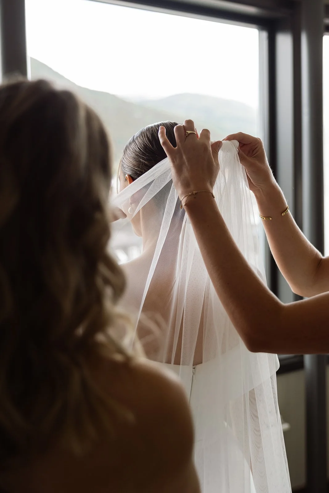 En kvinne hjelper en brud med å justere sløret foran et vindu. 