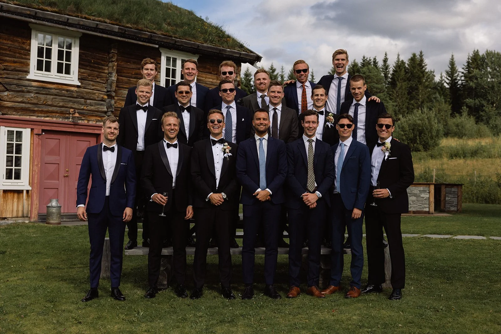 En gruppe menn i formelle dresser, noen med sløyfe, står foran en liten, trekledt hytte i et grønt utendørsområde. De er glade og smiler, noen holder drikke.