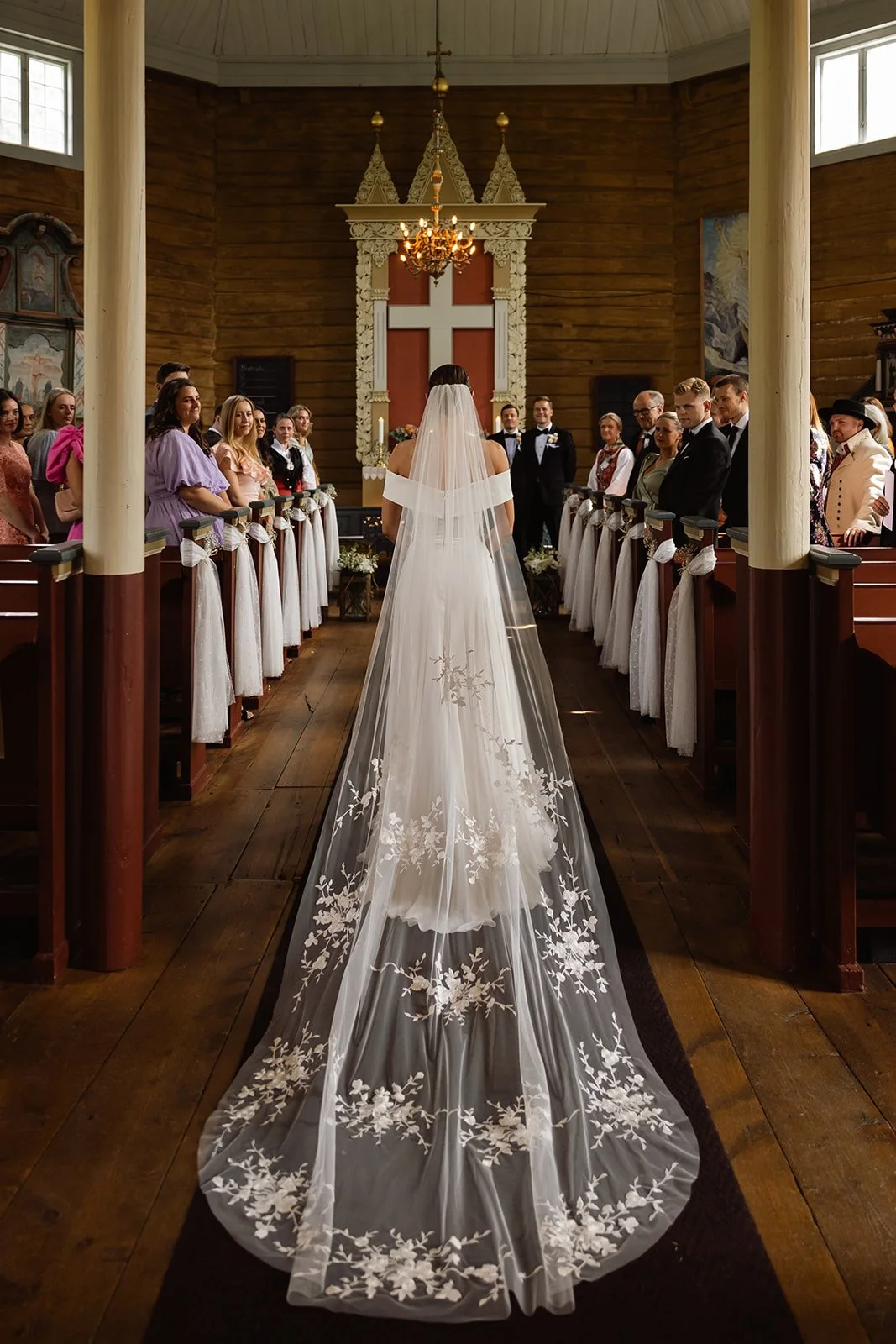 En brud i hvit kjole med langt, blomstret slør går ned midt i en kirke, omgitt av brudepiker og brudesvenner.