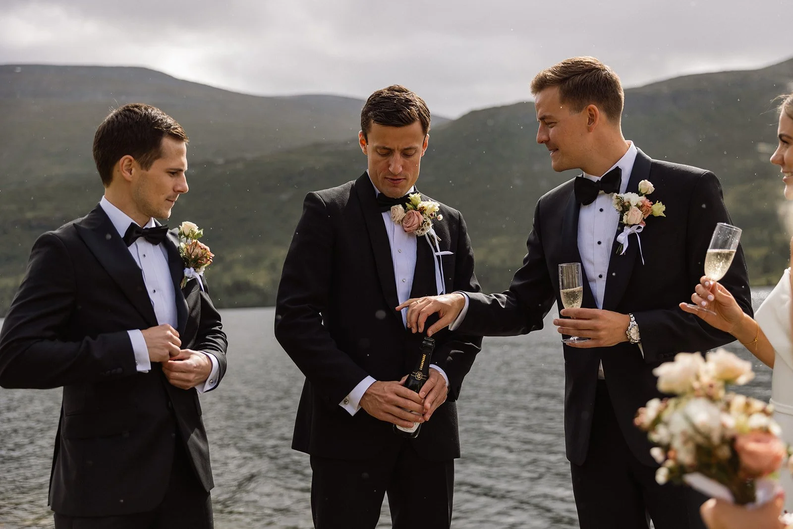 En gruppe gærne menn og en kvinne som holder en brudebukett under et bryllup i utendørsmiljø ved innsjø med fjell i bakgrunnen.