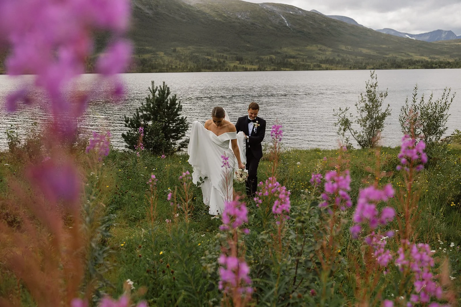 En brud og brudgom på bryllupsfotografering ved en innsjø omgitt av blomster og fjell