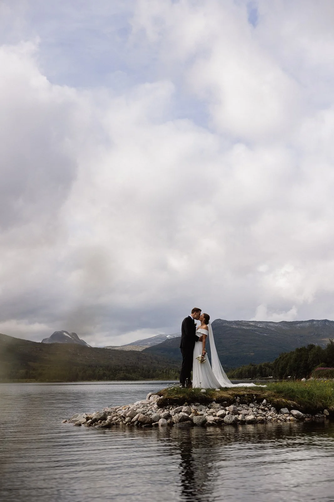 Et nygift par står på en liten holme i en innsjø, omgitt av fjell og skog, med overskyet himmel i bakgrunnen.