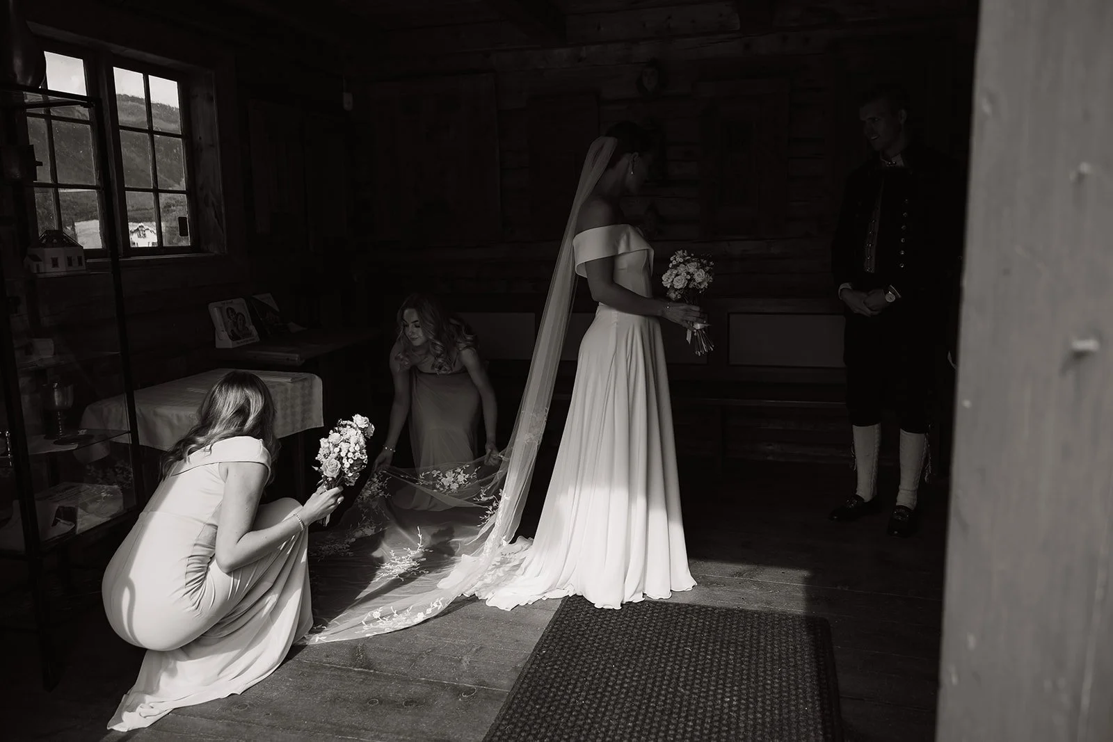 En bryllupsscene i et rom med tre kvinner og en mann. To kvinner hjelper bruden med en lang brudekjole og en bukett blomster, mens den tredje kvinnen står i bakgrunnen. Mannen står i hjørnet iført tradisjonell europeisk folkedrakt, smilete og ser på.