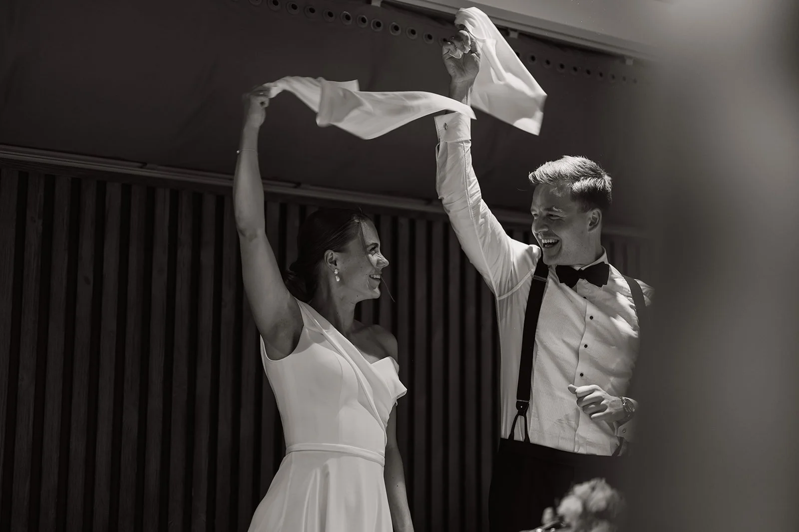 En lykkelig brudepar smiler og leker med et skjerf under en seremoni. De er kledd i formelle klær, og bakgrunnen er en vegg med vertikale treplanker.