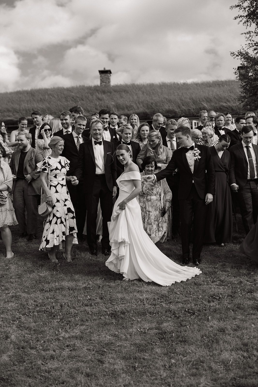 Sort-hvitt bilde av en bryllupsfeiring med bruden og brudgommen omgitt av gjester utendørs.