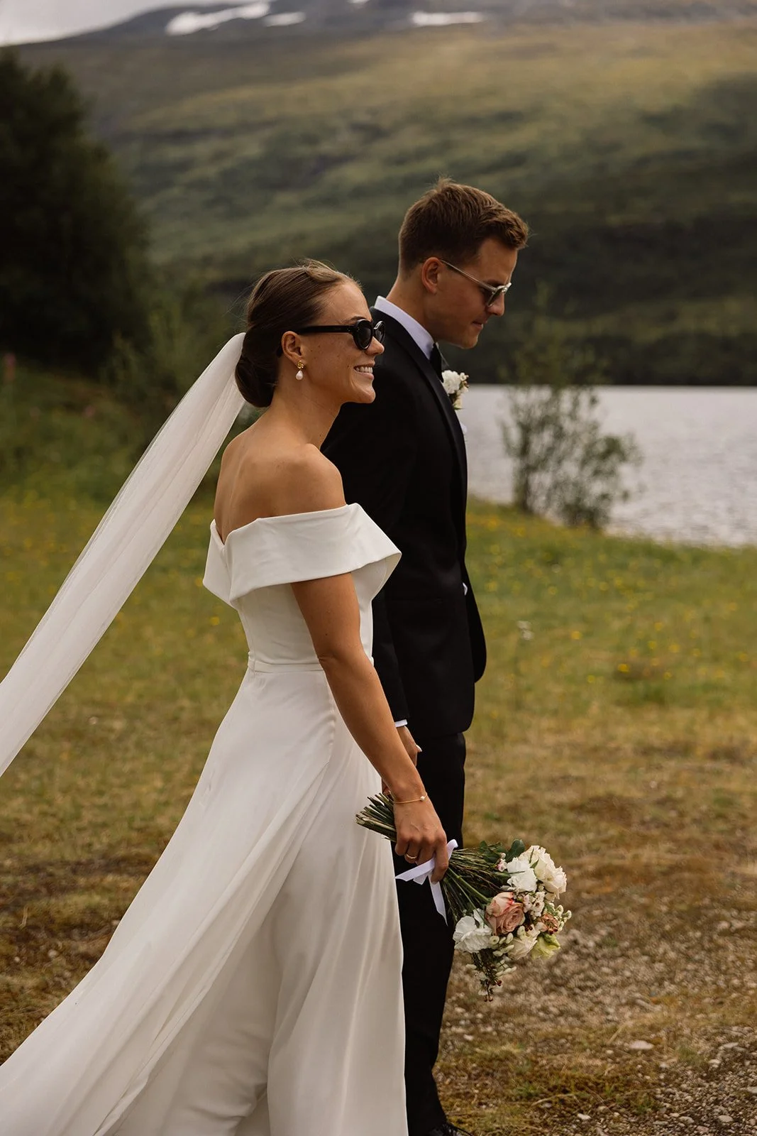 Et brudepar står ved en innsjø i fjellandskap, kvinnen holder en bukett med blomster og smiler, begge kledd i formell bryllupsantrekk, med fjell og vann i bakgrunnen.