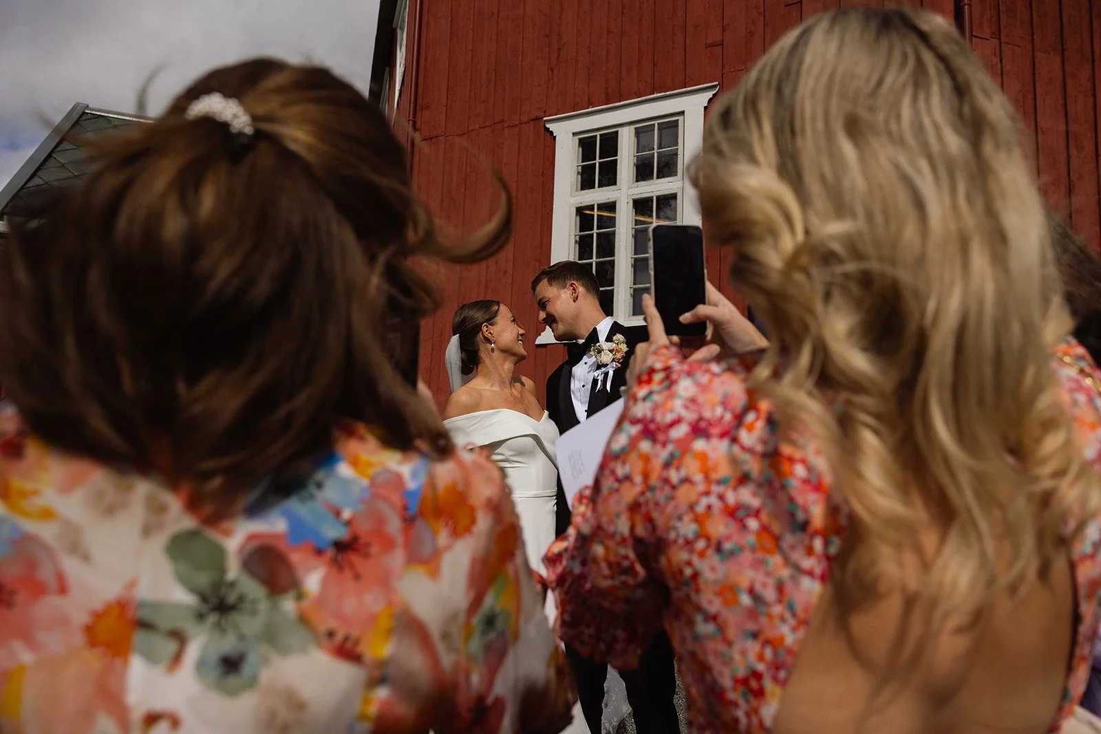 Et brudepar står nære hverandre og smiler til hverandre under et bryllup, omgitt av gjester. To kvinner i fargerike blomsterkjoler tar bilder av paret, mens en person holder en papir, på en utvendig rød vegg.