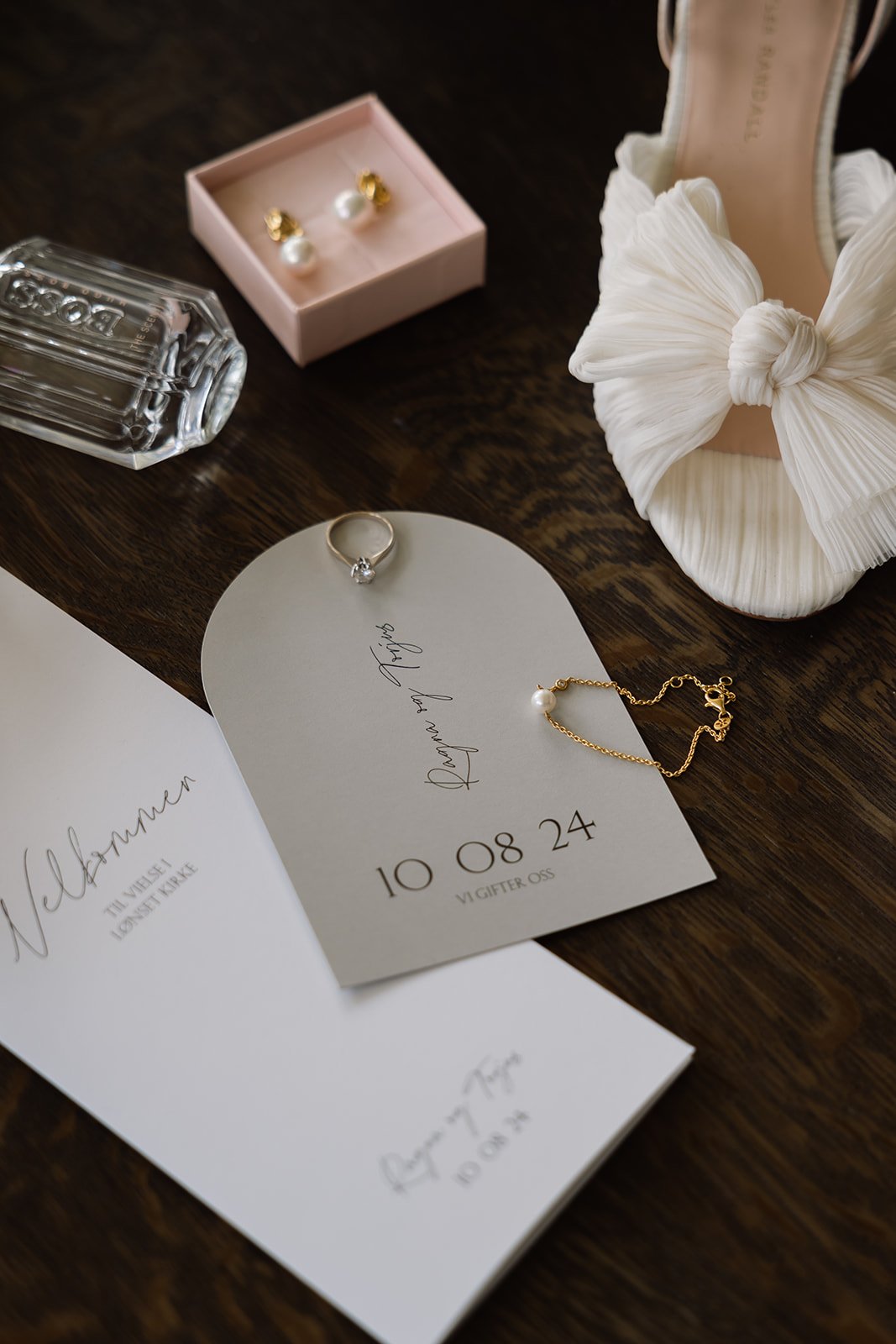Bryllupsdetaljer inkludert et diamantringer, perlesmykker og et bryllupsinvitasjon med datoen 10.08.24.