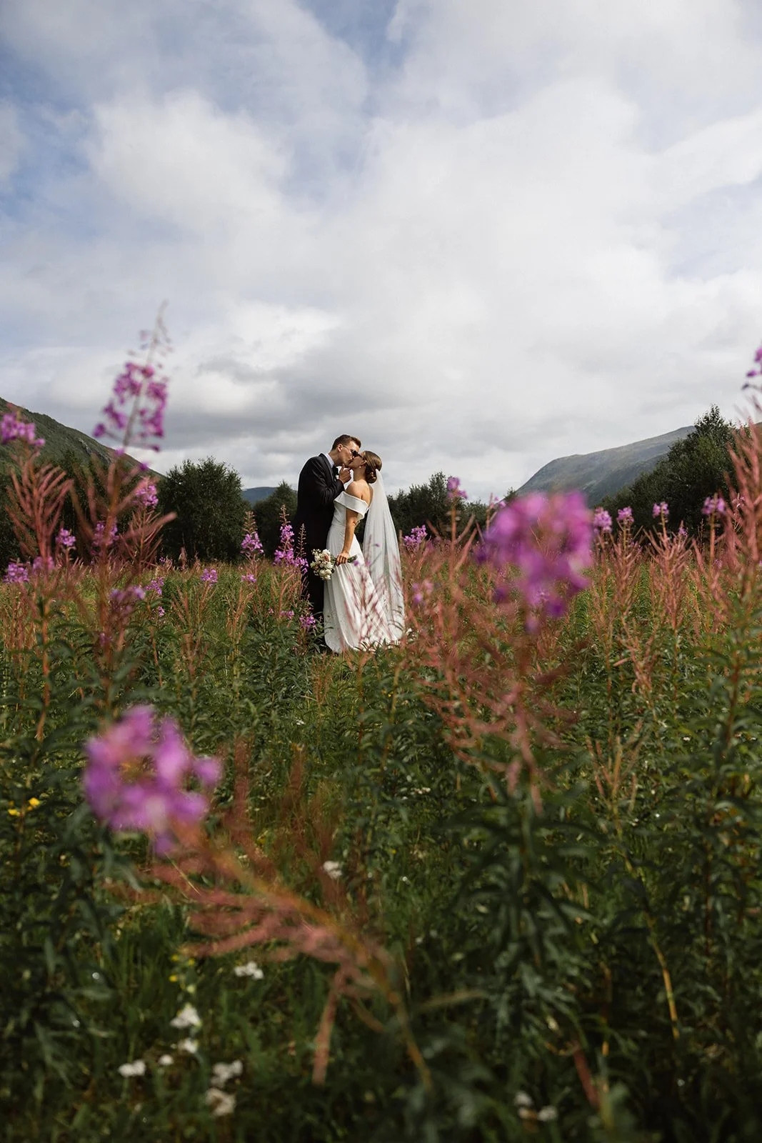 Et brudepar som kysser i en blomstereng med fjell og skyer i bakgrunnen.