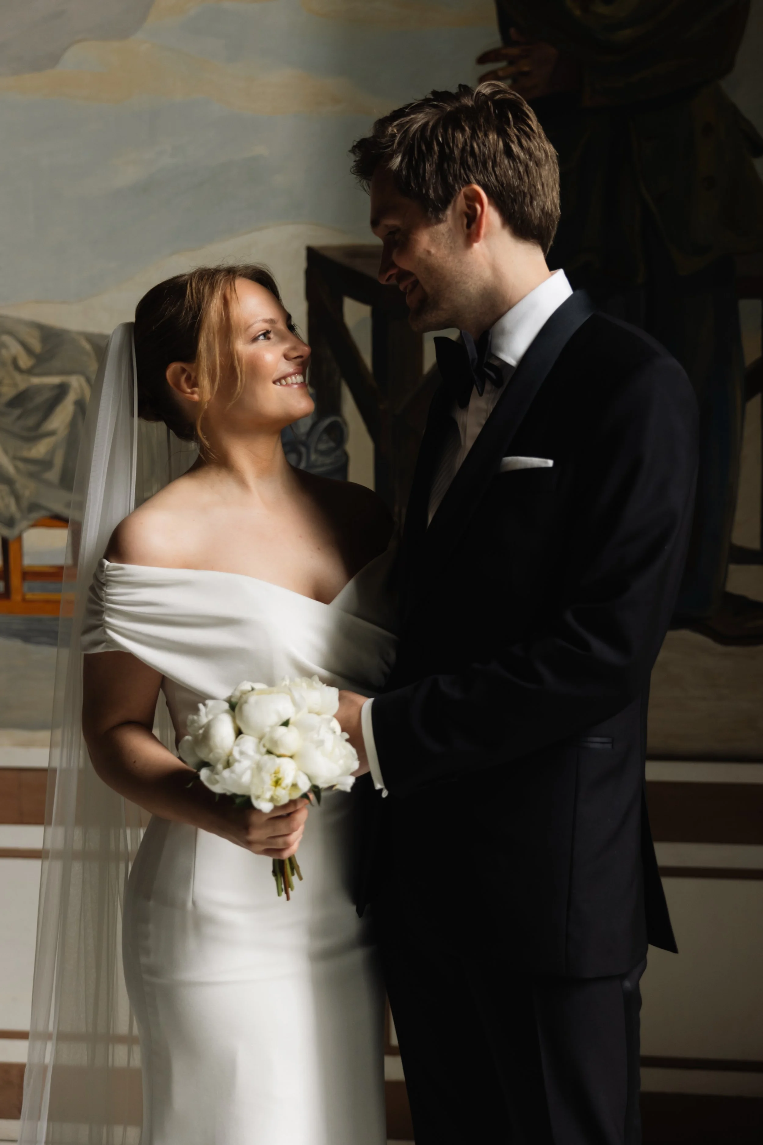 Et bryllupsbilde av et brudepar som smiler til hverandre, med bruden som holder en blomsterbukett, innendørs.