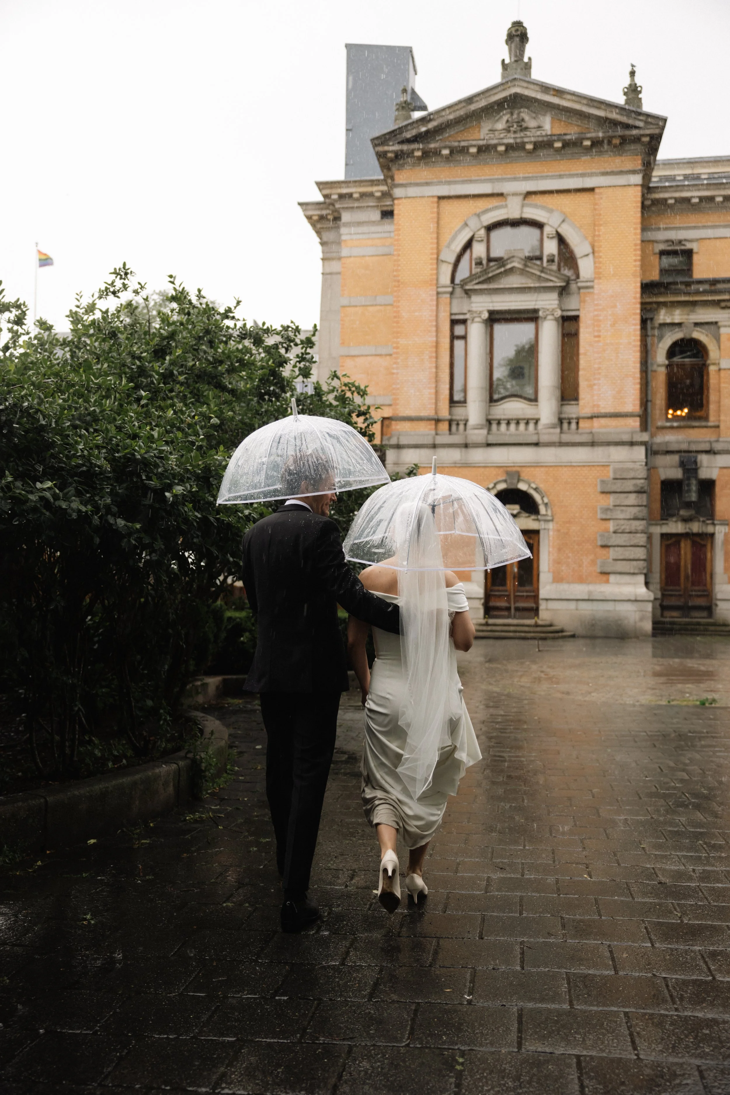 En brud og en brudgom går under paraplyer foran en gammel bygning på en regnfull dag.