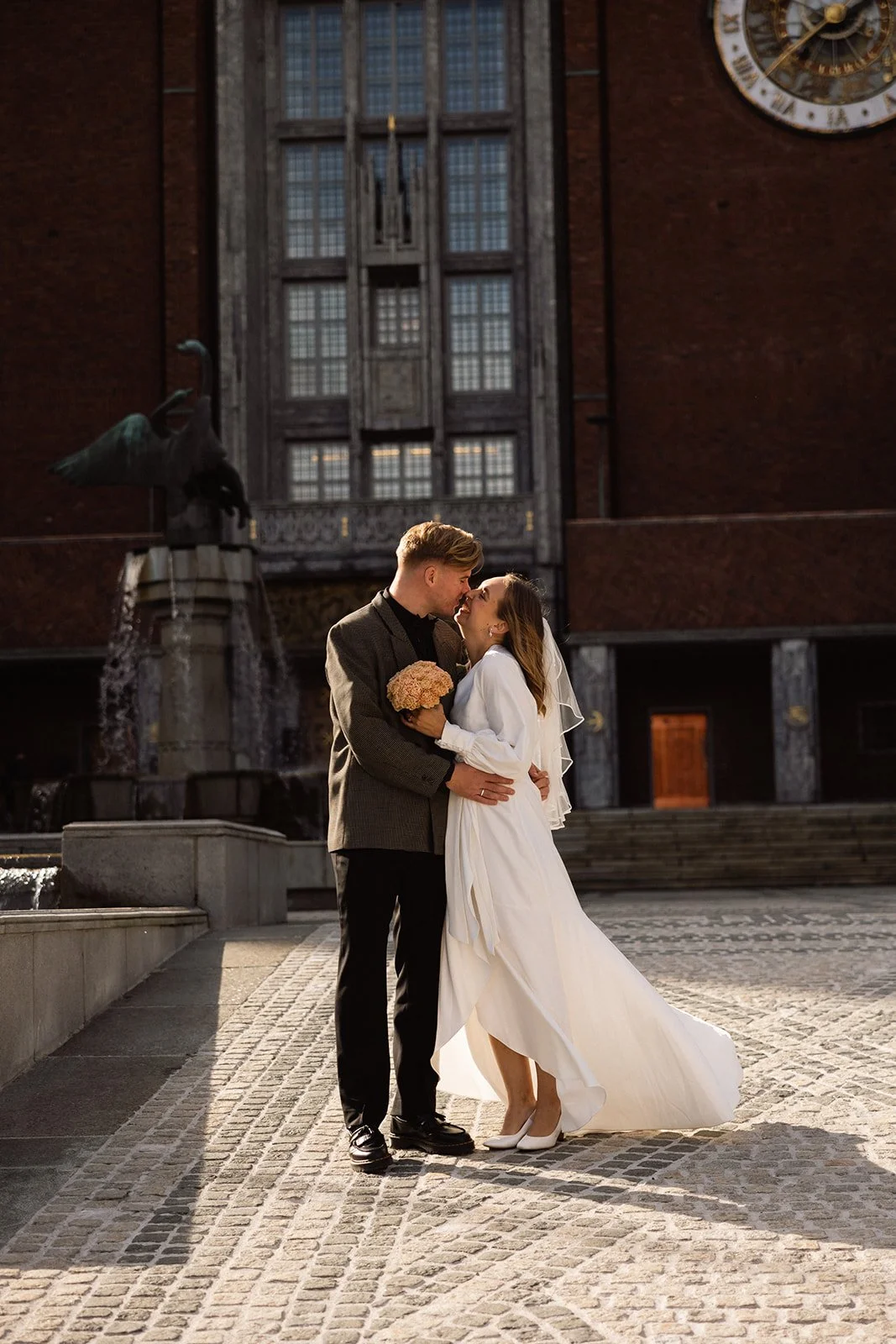 Et nytt ektepar står tett sammen foran en vannfontene og en stor bygning med ruter i vinduene i bakgrunnen.
