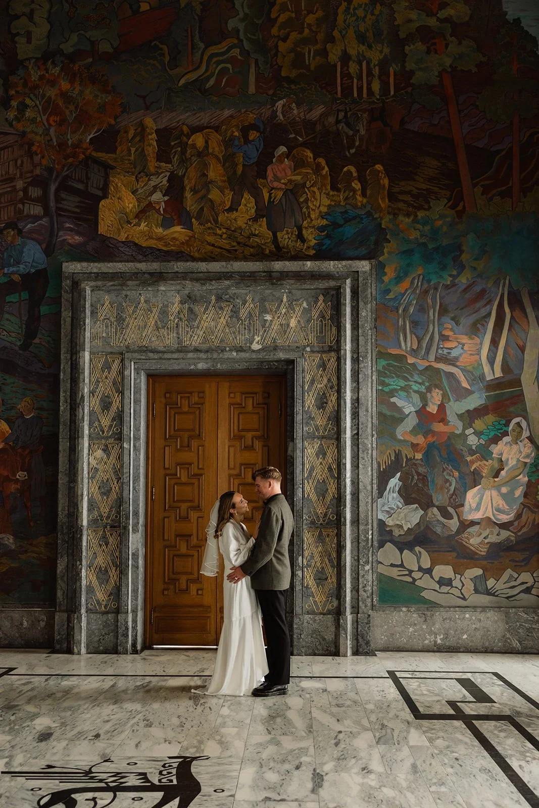 Et nygift par står og ser hverandre dypt i øynene, i en stor hall med et malt veggmaleri som skildrer tradisjonelt arbete, natur og folk i en norsk landsby, med en trapp og marmorgulv.