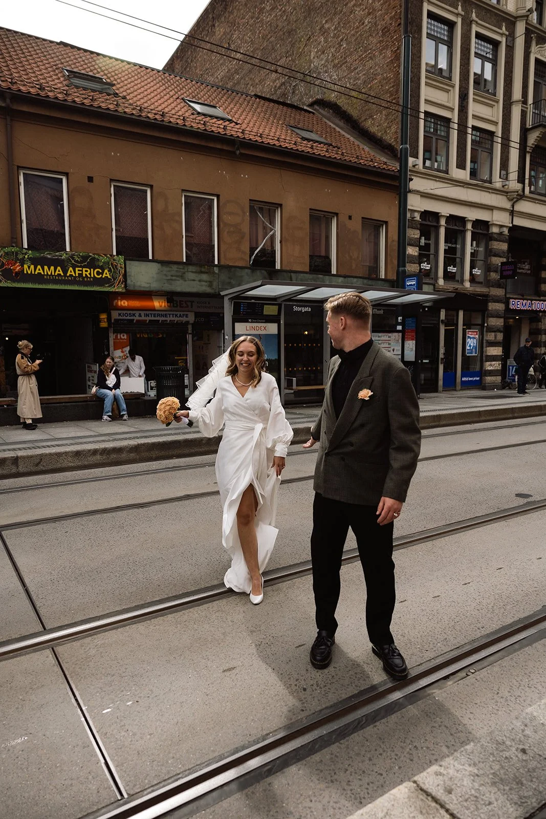 En nygift brud og brudgom går sammen på en bygate med togskinner, med bygninger og busstoppesteder i bakgrunnen.
