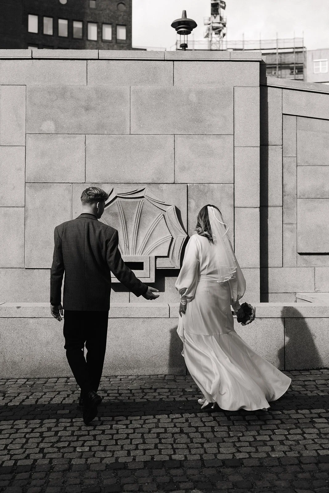 To personer, en mann i dress og en kvinn i brudekjole, går sammen på brostein, ved en vegg med et kunstnerisk innrammet utskjæring. Kvinn har brudekrone og holding en bukett, de ser ut til å være på vei til en bryllupsseremoni.