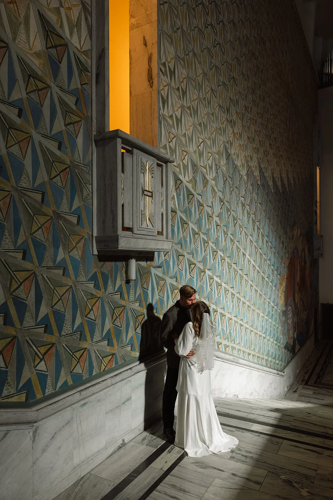 Et par i formelle klær står nær hverandre foran en dekorert vegg i en stor hall, med en fløyelsklut i hvitt.
