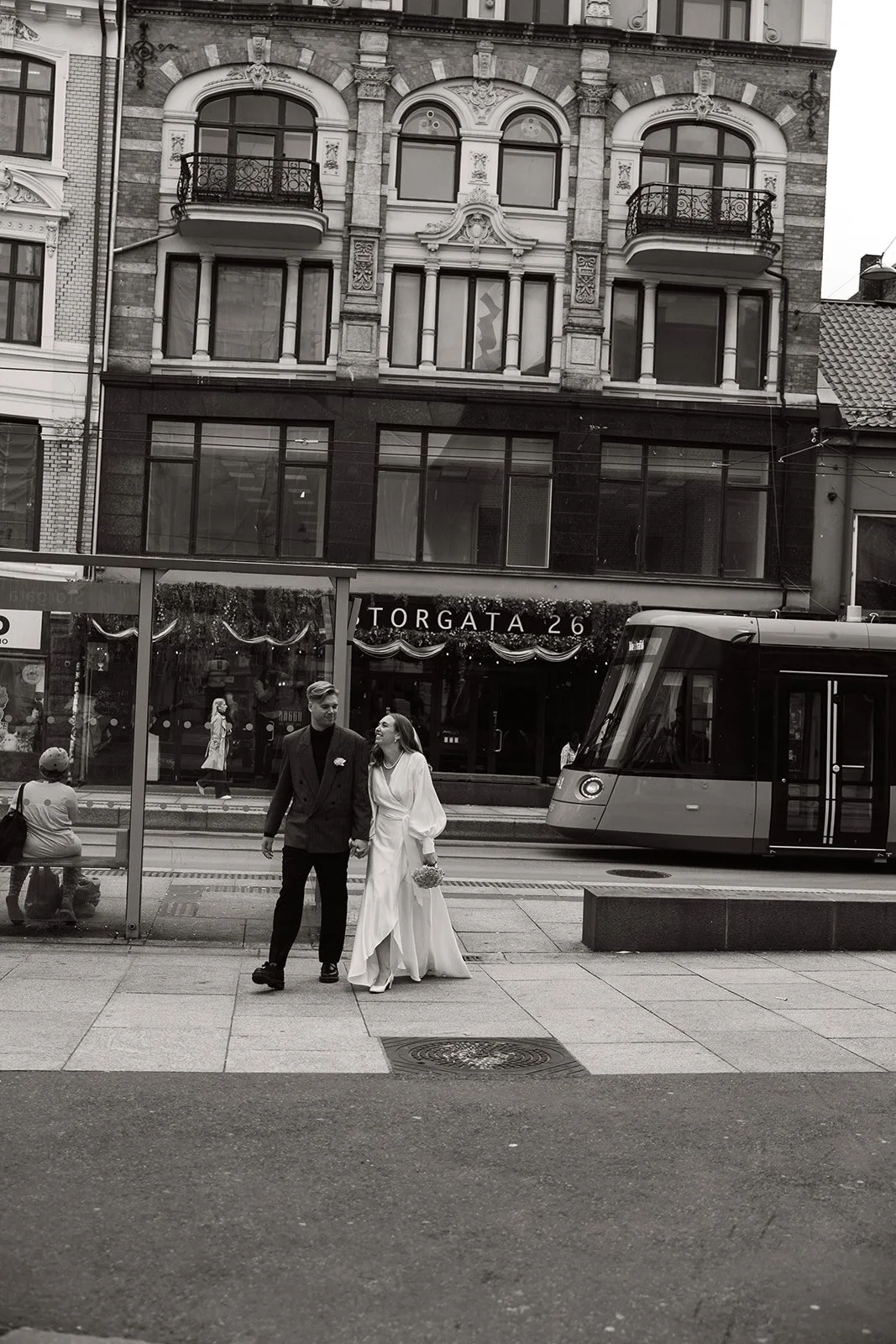 To personer i brudekjole og dress går hånd i hånd på en fortauskafé, med en trikk og et gammelt bygning i bakgrunnen, på en bygate.