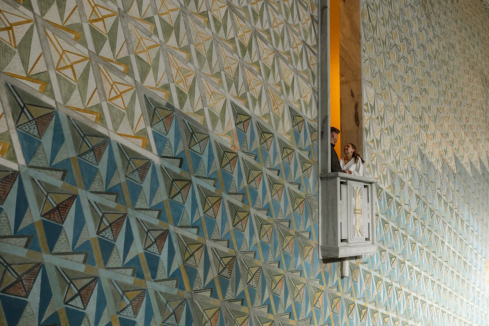 To personer i en liten balkong, omgitt av en stor vegg med geometrisk mønster i grønne, blå, gule og grå farger.