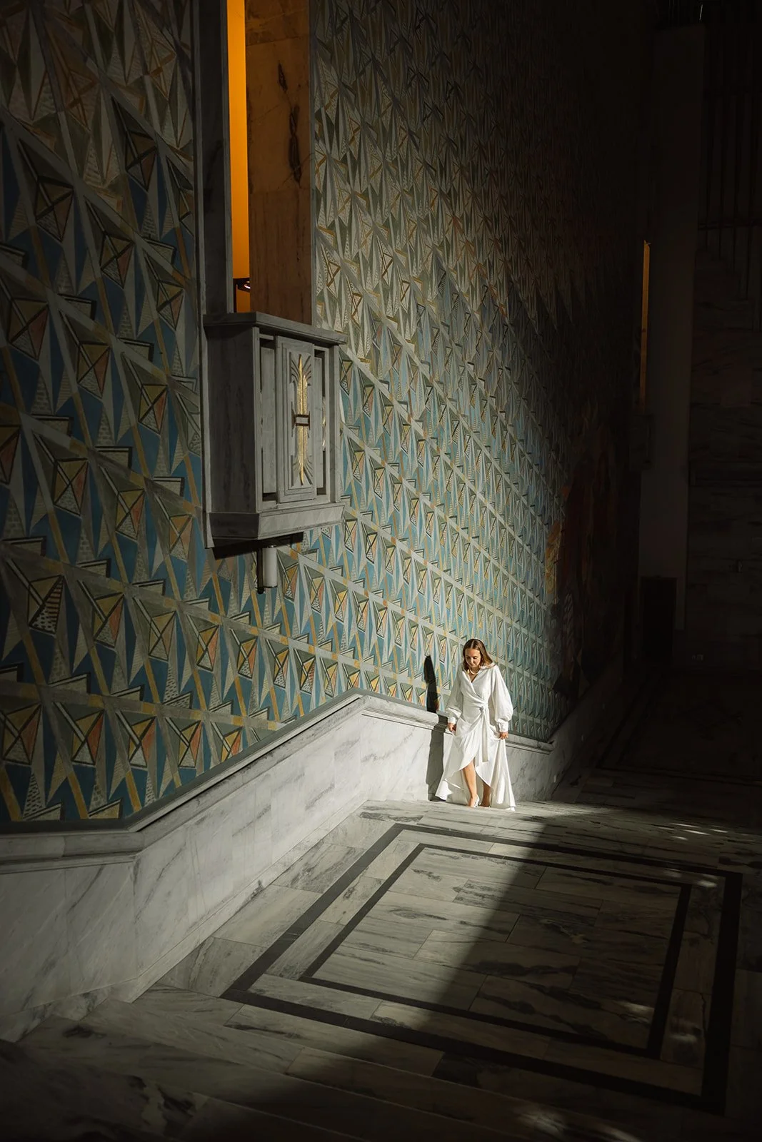 En kvinne i hvit kjole går opp en trapp i et rom med mønstret vegg og marmorgulv, opplyst av naturlig lys.