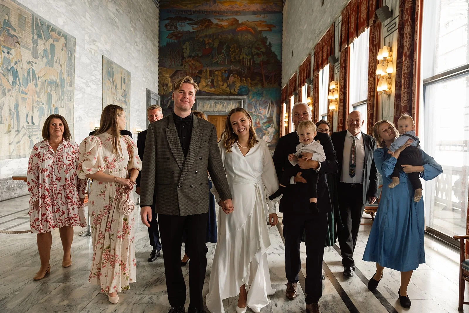 En gruppe mennesker i formelle klær, etpar i midten holder hender, inne i et rom med store vinduer og dekorative gardiner, i bakgrunnen et stort kunstverk på veggen.