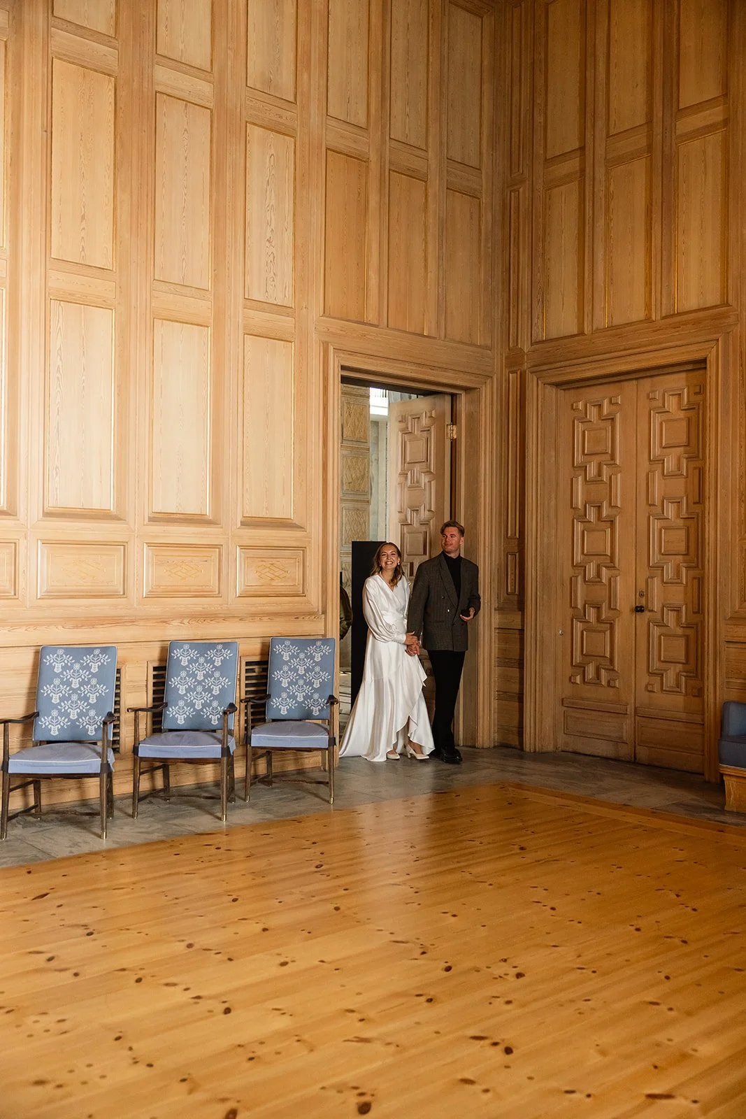 To personer står i en stor, veggdekket rom med tregrønn på gulvet, kledd i elegante klær, og smiler.