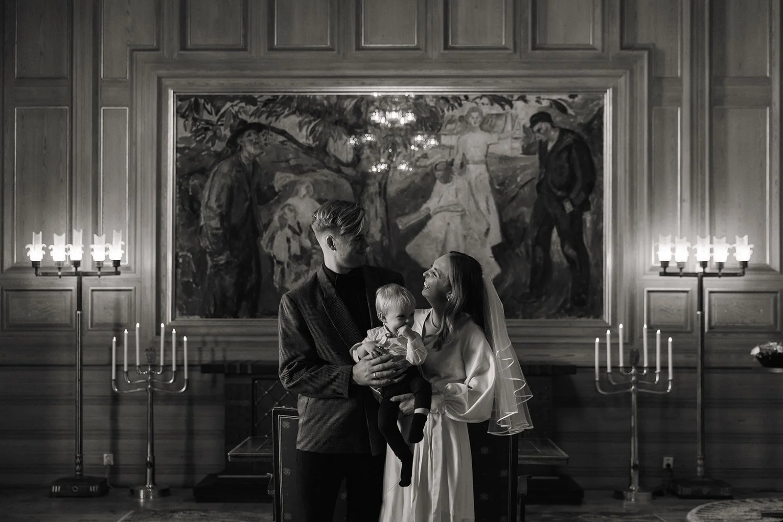 Et ungt par og en kvinne med en liten jente står sammen i et kirkerom, alle smiler. Parret står foran en stor vegg med et maleri, lysende lysestaker er plassert på hver side.