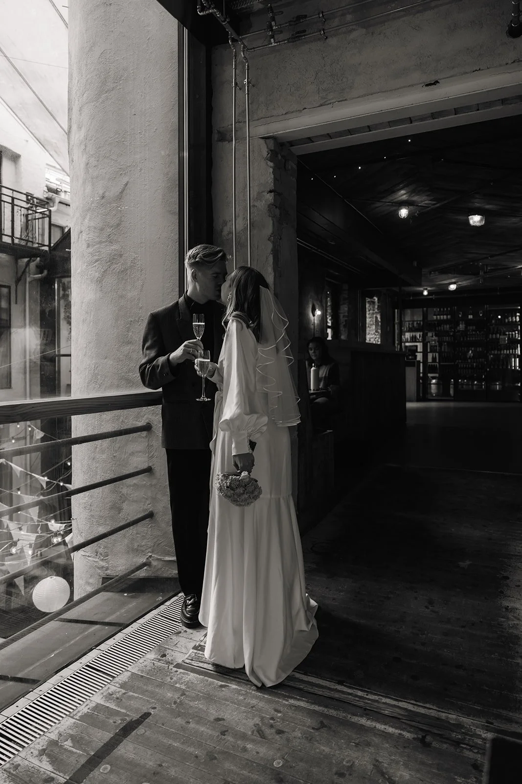 Et bilde av et brudepar som står nært ved et vindu, deler et kyss mens de holder champagneglass, i en mørk innendørs setting.