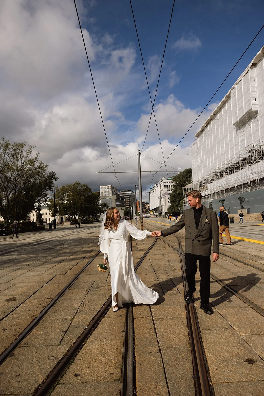 To personer, en kvinne i en lang, hvit brudekjole og en mann i en grå dress, holder hender på et byområde med sporvogner, bygninger og noen trær i bakgrunnen.