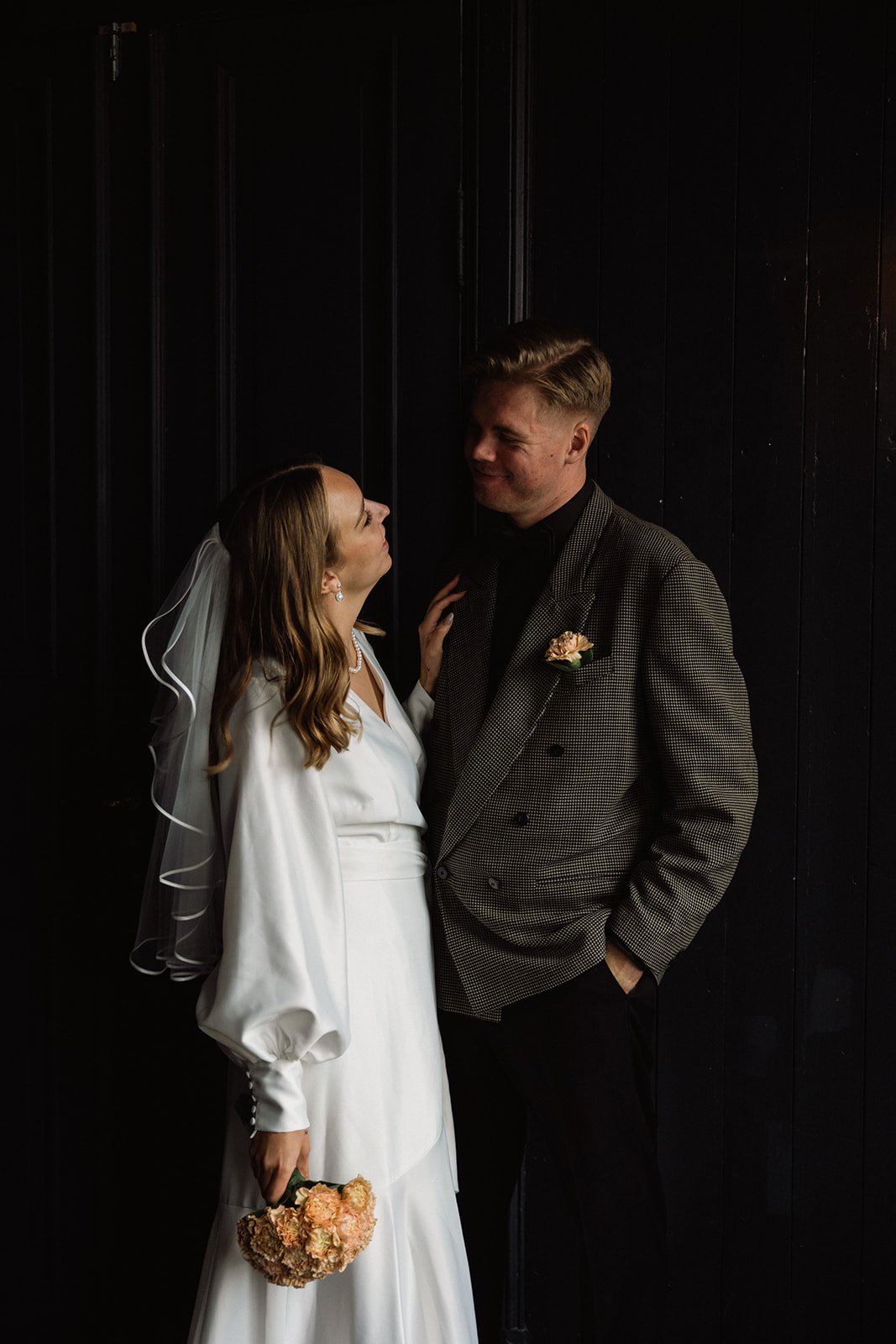 Et brudepar står tett sammen i et rom med mørke vegger. Bruden har på seg en hvit kjole med puffede ermer og holder en bukett med lyse blomster. Mannen har på seg en rutete dressjakke med sort skjorte og har et blomsterarrangement i brystlommen. De s