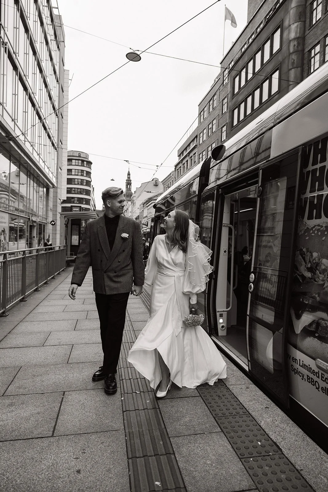 To personer, en mann og en kvinne i brudeklær, går hånd i hånd ut av en sporvogn i en bygate. Kvinnen holder en liten kurv og har på seg en hvit brudekjole og brudebryllupslue, mens mannen har på seg en mørk dress. Bildet er i svart-hvitt.
