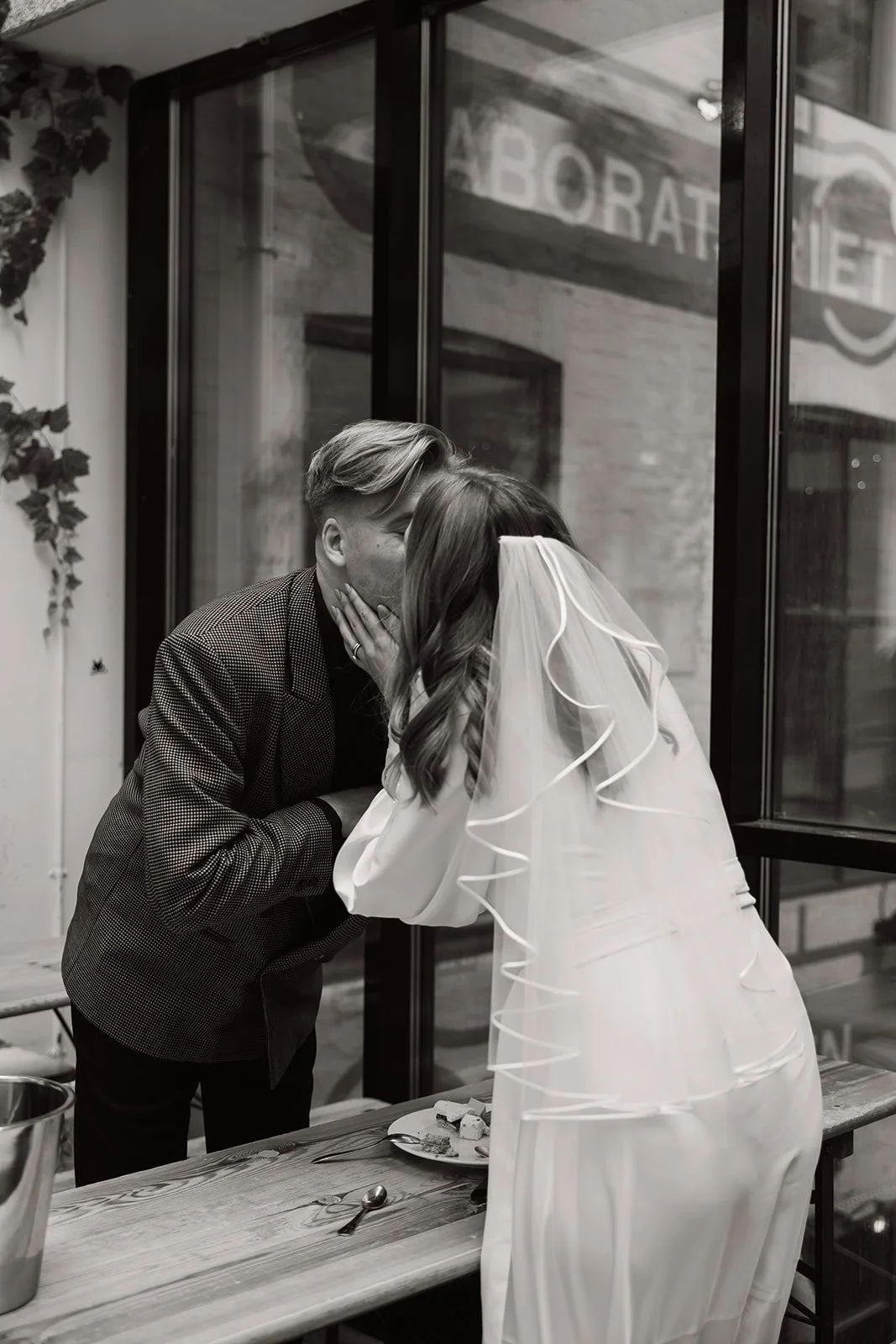 Bruden og mannen kysser hverandre på bryllupsfeiring, med bryllupsdrakt og påkledning, inne i et rom med store vinduer og trebord.