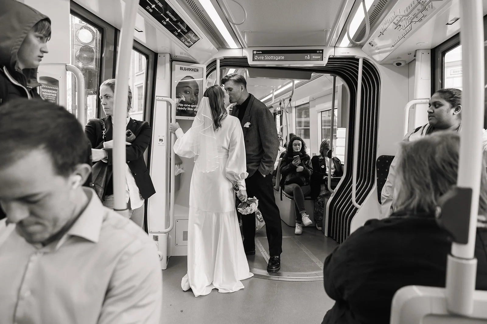 To personer i brudekjole og dress står nær hverandre i en trikk, omgitt av passasjerer som sitter og står. Flere passasjerer ser på sine telefoner, noen ser bort, og noen vender blikket mot de som står. Det er en reklameskilt og en digital informasjo