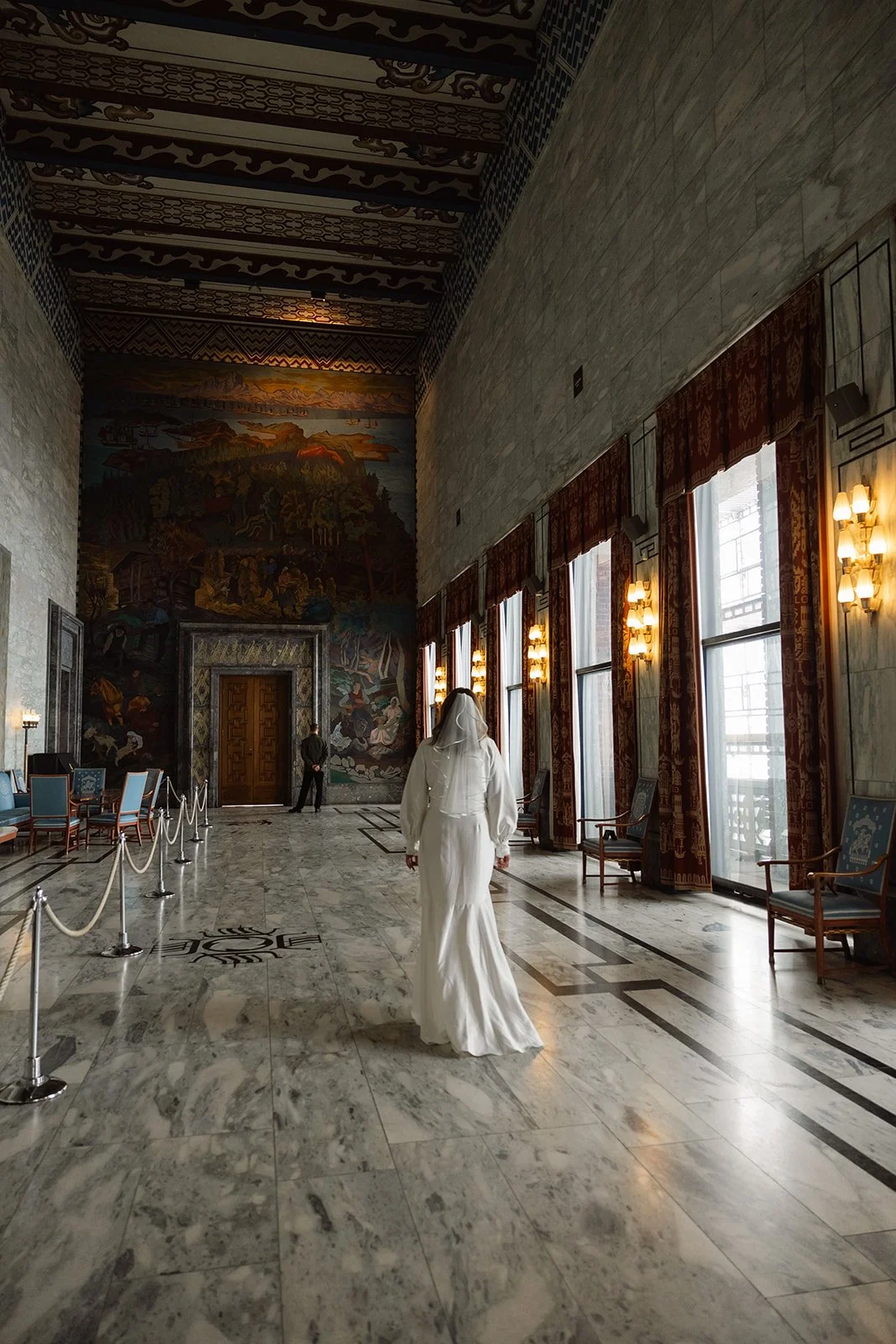 En kvinne i brudekjole som går inn i et stort og elegant rom med store vinduer, marmorgulv og lyse gardiner. Veggen bak har en stor, fargerik veggmaleri.