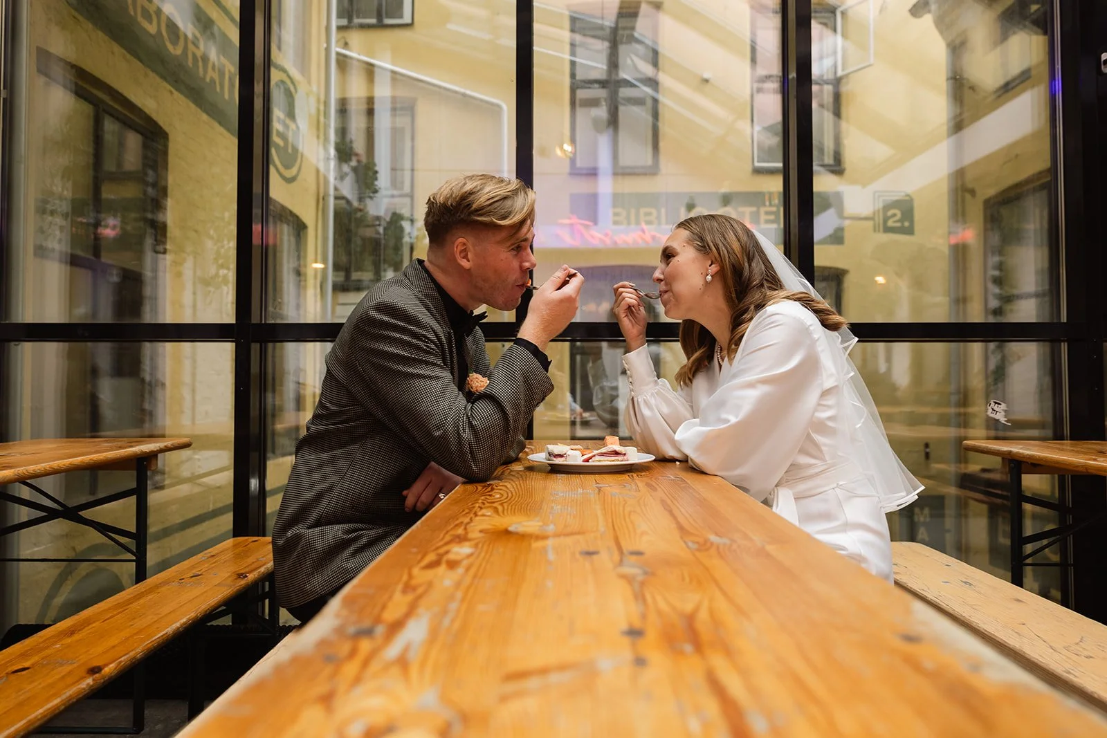 Et ungt par som kler seg i bryllupsklær, sitter ved et langt trebord på en kafé og deler en dessert.
